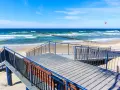 Drewniany deptak prowadzi do plaży w Grzybowie. W oddali widać morze, falochrony i kitesurferów. To idealne miejsce na relaks i podziwianie atrakcji nadmorskich.