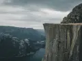 Człowiek w żółtej kurtce stoi na krawędzi skalnego klifu w Norwegii, podziwiając majestatyczny fiord i góry.