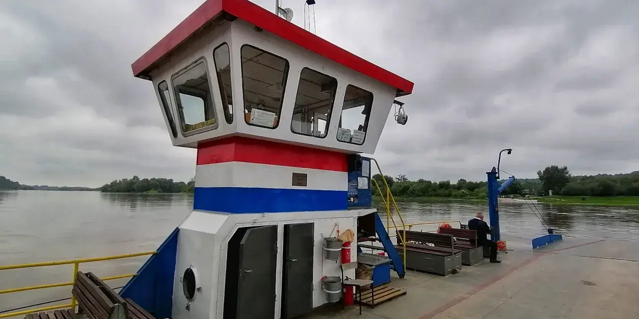 Prom z pomostem, kabiną kapitańską i ławkami na tle rzeki i zachmurzonego nieba.