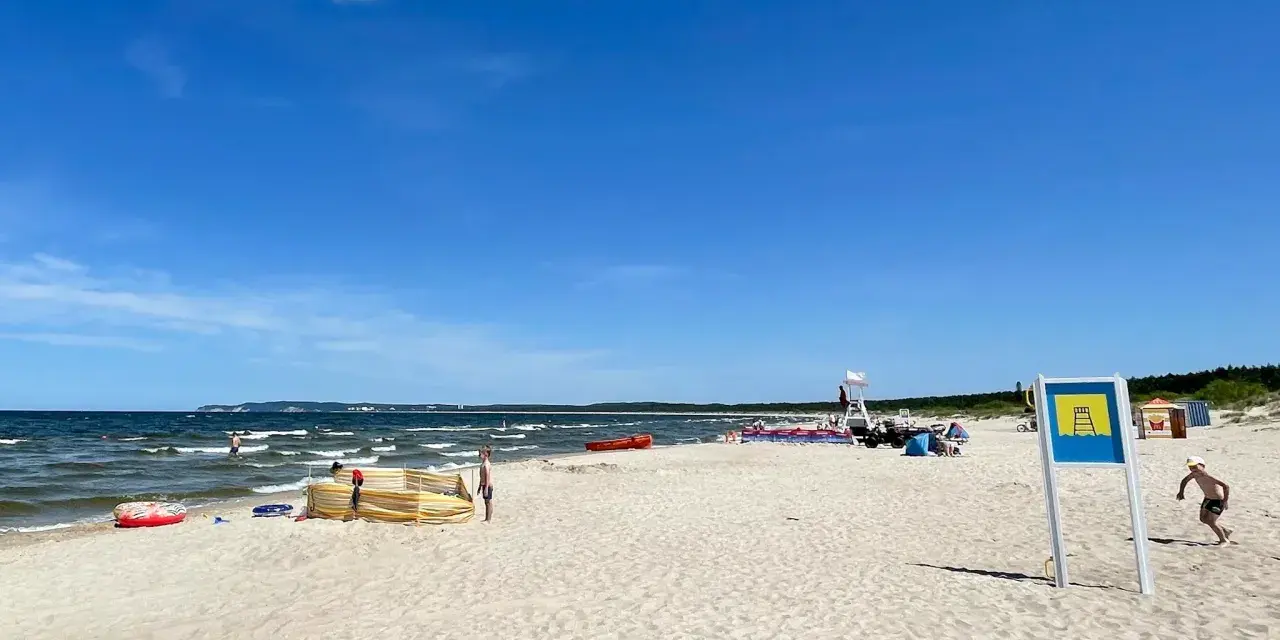 Słoneczna plaża Cztery Wiatry z piaskiem, morzem i ludźmi. W oddali widać ratownika na wieży.
