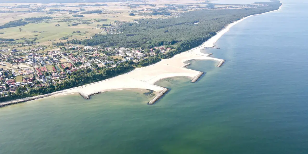 Jarosławiec nad morzem: szeroka, piaszczysta plaża z kamiennymi falochronami, otoczona lasem sosnowym i zabudową letniskową.