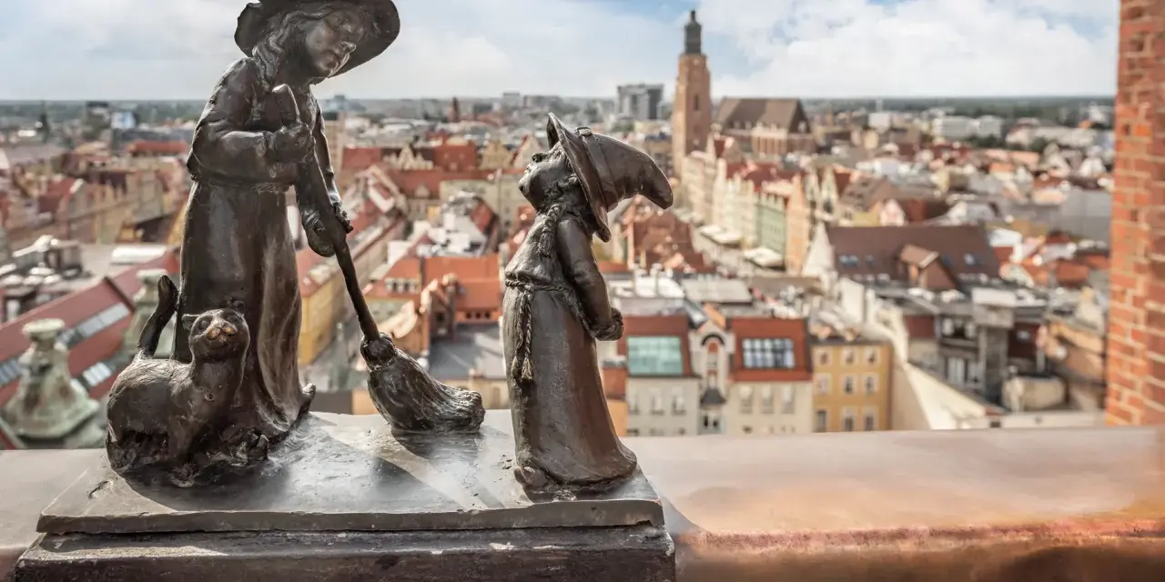 Figurki krasnali: czarownica z kotem i miotłą, na tle panoramy Wrocławia.