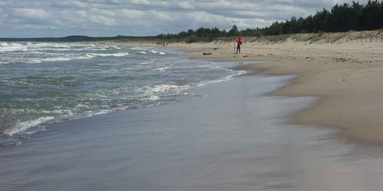 Plaża z falującym morzem i osobą w czerwonej bluzie na piasku.