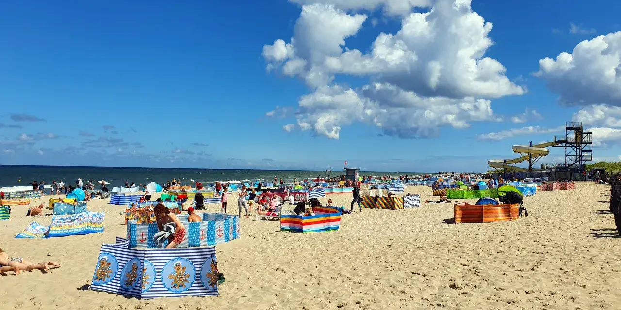 Władysławowo z dzieckiem: plaża pełna kolorowych parawanów, ludzi odpoczywających i bawiących się na piasku, w tle morze i zjeżdżalnie wodne.