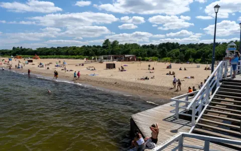 Słoneczny dzień nad morzem bałtyckim. Ludzie odpoczywają na piaszczystej plaży, niektórzy kąpią się w wodzie. Drewniane molo prowadzi na plażę.