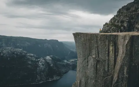 Człowiek w żółtej kurtce stoi na krawędzi skalnego klifu w Norwegii, podziwiając majestatyczny fiord i góry.