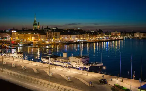 Wieczorny Sztokholm w jeden dzień: oświetlone budynki, odbijające się w wodzie światła, statek przy nabrzeżu.