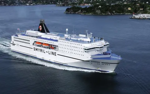 Duży biały prom "Smyril-Line Norrona" płynie po spokojnym morzu, w tle widać zielone wybrzeże.