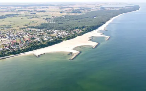Jarosławiec nad morzem: szeroka, piaszczysta plaża z kamiennymi falochronami, otoczona lasem sosnowym i zabudową letniskową.