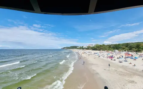 Słoneczna plaża z tłumem turystów, gdzie jest najcieplejsze morze. Woda faluje, a na piasku rozłożone są parawany i ręczniki.