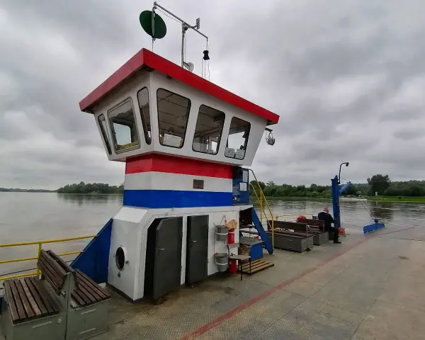 Prom z pomostem, kabiną kapitańską i ławkami na tle rzeki i zachmurzonego nieba.