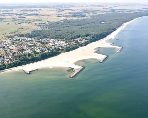Jarosławiec nad morzem: szeroka, piaszczysta plaża z kamiennymi falochronami, otoczona lasem sosnowym i zabudową letniskową.