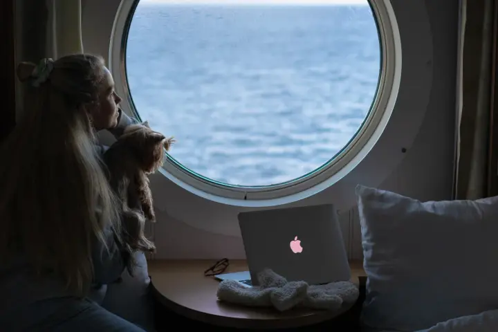Dziewczyna z psem patrzy przez okrągłe okno na morze. Obok leży laptop z podświetlonym logo, jak prom nova star.