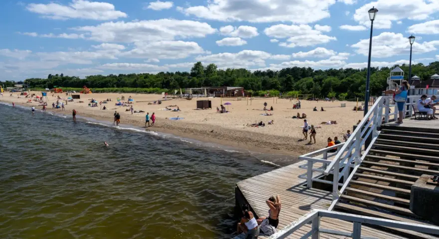 Słoneczny dzień nad morzem bałtyckim. Ludzie odpoczywają na piaszczystej plaży, niektórzy kąpią się w wodzie. Drewniane molo prowadzi na plażę.