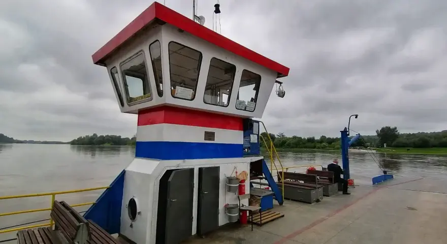 Prom z pomostem, kabiną kapitańską i ławkami na tle rzeki i zachmurzonego nieba.