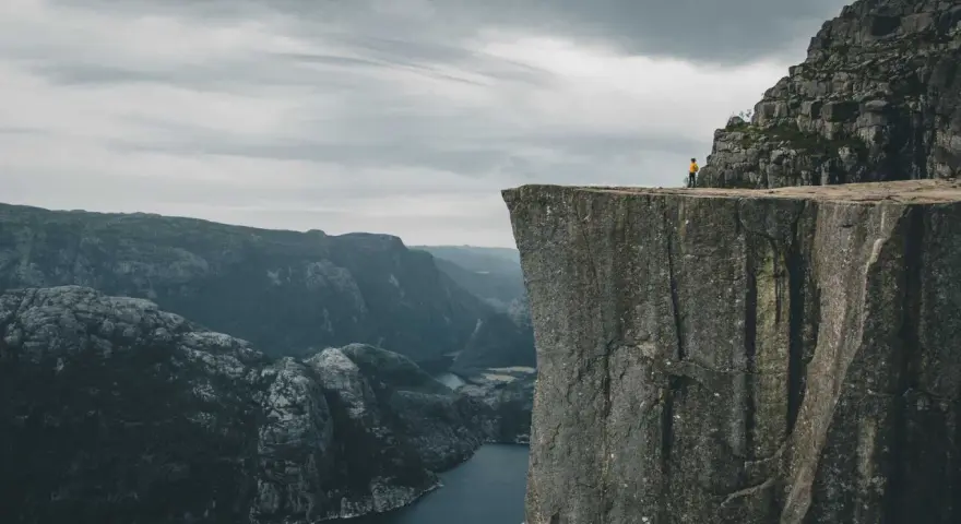 Człowiek w żółtej kurtce stoi na krawędzi skalnego klifu w Norwegii, podziwiając majestatyczny fiord i góry.