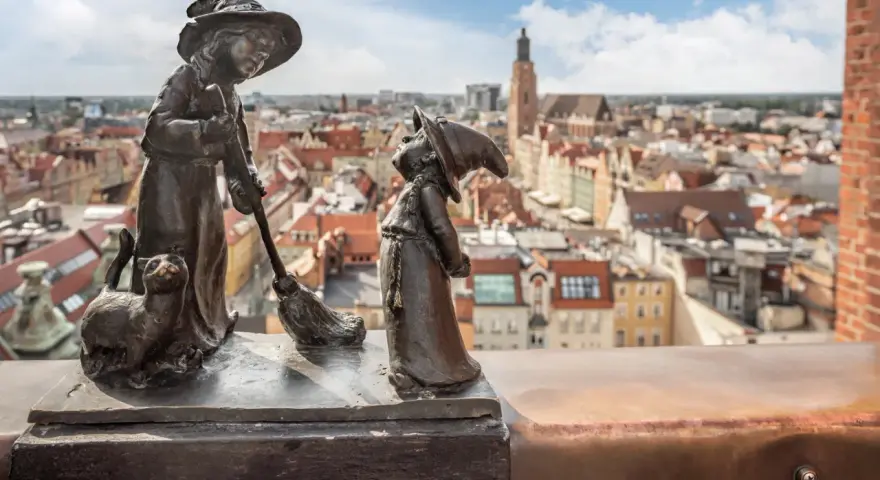 Figurki krasnali: czarownica z kotem i miotłą, na tle panoramy Wrocławia.