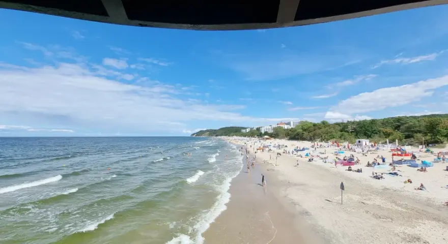 Słoneczna plaża z tłumem turystów, gdzie jest najcieplejsze morze. Woda faluje, a na piasku rozłożone są parawany i ręczniki.