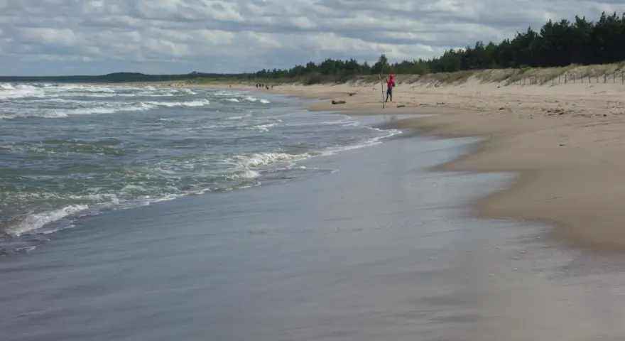 Plaża z falującym morzem i osobą w czerwonej bluzie na piasku.