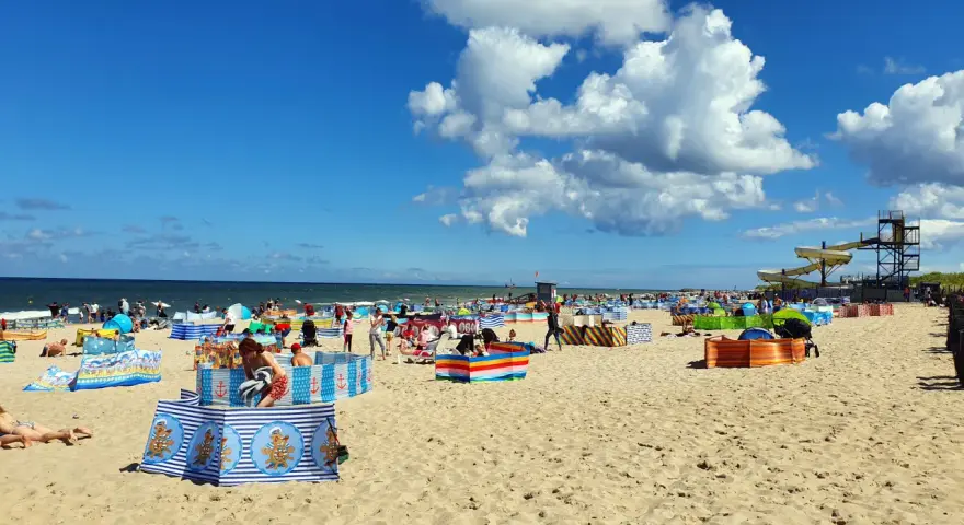 Władysławowo z dzieckiem: plaża pełna kolorowych parawanów, ludzi odpoczywających i bawiących się na piasku, w tle morze i zjeżdżalnie wodne.