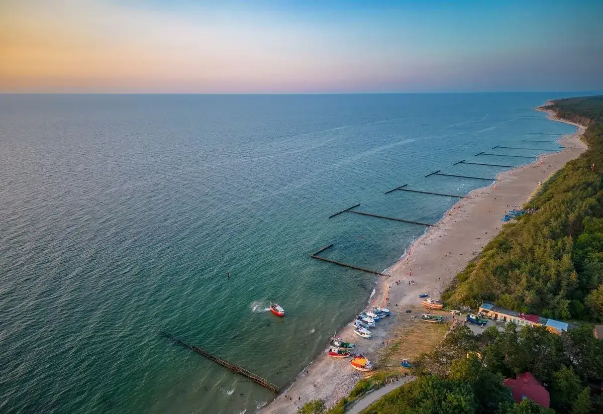 Piękny widok na plażę w Jarosławcu nad morzem. Woda jest spokojna, a na brzegu widać kilka łodzi i spacerujących ludzi.