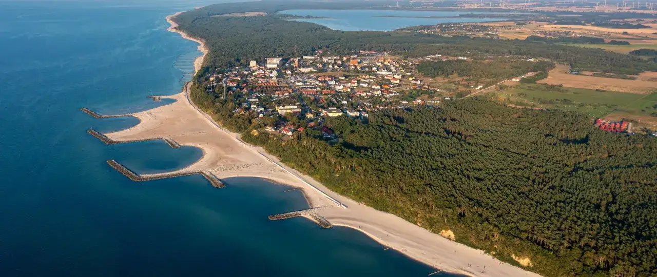 Jarosławiec nad morzem, z piaszczystą plażą, falochronami i lasem sosnowym otaczającym zabudowania.