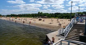 Słoneczny dzień nad morzem bałtyckim. Ludzie odpoczywają na piaszczystej plaży, niektórzy kąpią się w wodzie. Drewniane molo prowadzi na plażę.