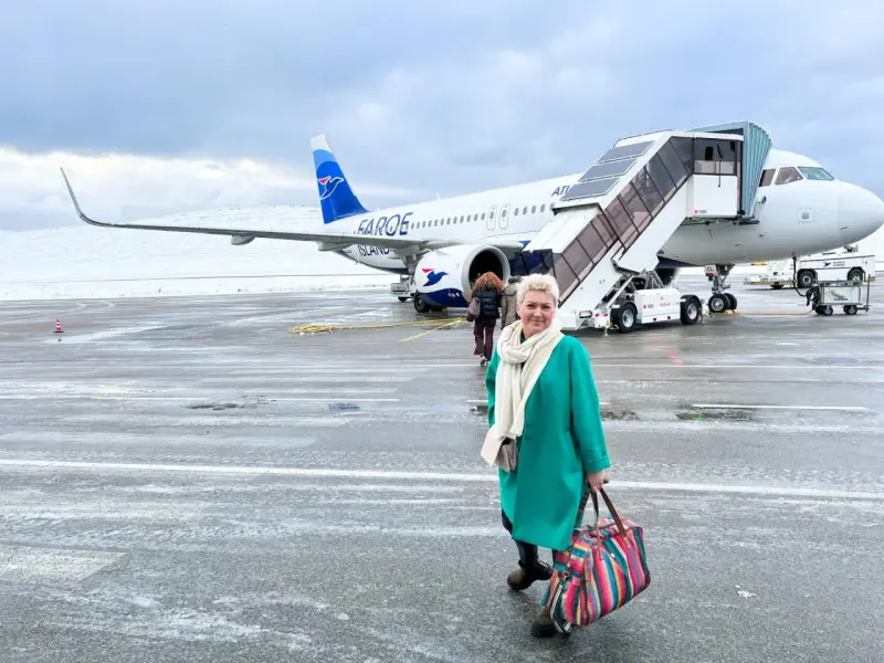 Kobieta z kolorową torbą na lotnisku, gotowa do podróży. Samolot Atlantic Airways z napisem "Faroe Islands" sugeruje, jak dostać się na Wyspy Owcze.